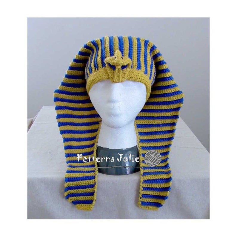 K&ouml;nnte beinhalten: Eine geh&auml;kelte M&uuml;tze mit einem blau-gelben Streifenmuster, die an die Kopfbedeckung eines &auml;gyptischen Pharaos erinnert. Die M&uuml;tze wird auf einem wei&szlig;en Mannequin-Kopf pr&auml;sentiert. Der Text "Patterns Jolie" ist auf dem Sockel des Mannequins sichtbar.