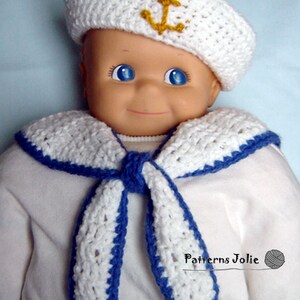 May include: A white crocheted sailor hat with a gold anchor on top, and a white crocheted sailor collar with blue trim. The hat and collar are worn by a doll.