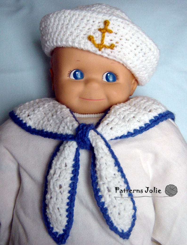 May include: A white crocheted sailor hat with a gold anchor on top, and a white crocheted sailor collar with blue trim. The hat and collar are worn by a doll.