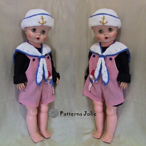 May include: Two dolls dressed in sailor outfits. Both dolls are wearing white sailor hats with a gold anchor, a white crocheted sailor collar, a blue and white striped shirt, and pink overalls. The dolls are standing on a beige surface.