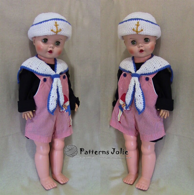 May include: Two dolls dressed in sailor outfits. Both dolls are wearing white sailor hats with a gold anchor, a white crocheted sailor collar, a blue and white striped shirt, and pink overalls. The dolls are standing on a beige surface.