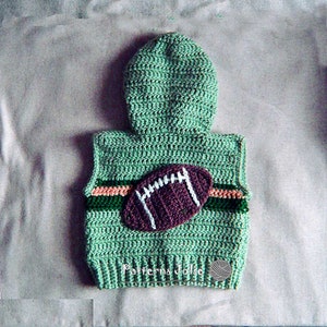 以下が含まれることがあります： フード付きの緑色の編み物ベストと茶色のフットボールのアップリケ。ベストは真ん中に緑、ピンク、白のストライプのバンドが付いています。ベストの下部に「Patterns Jolie」というテキストが表示されています。