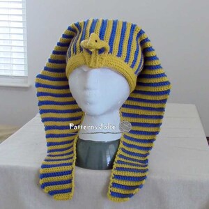 K&ouml;nnte beinhalten: Eine gestrickte M&uuml;tze mit gelben und blauen Streifen und einer kleinen, gelben, geh&auml;kelten Figur auf der Oberseite. Die M&uuml;tze ist in Form eines &auml;gyptischen Pharaonenkopfschmucks geformt.