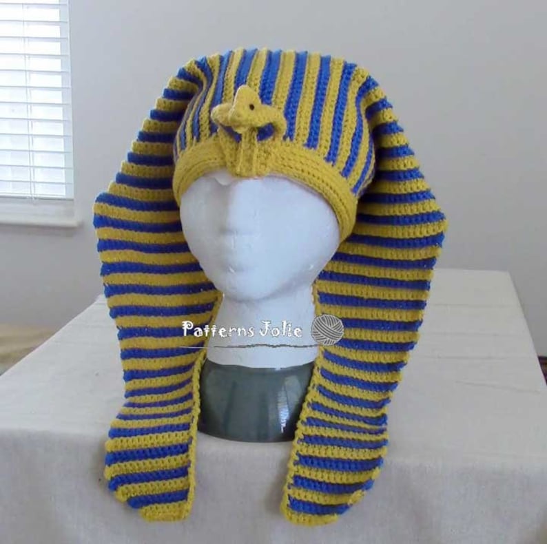 K&ouml;nnte beinhalten: Eine gestrickte M&uuml;tze mit gelben und blauen Streifen und einer kleinen, gelben, geh&auml;kelten Figur auf der Oberseite. Die M&uuml;tze ist in Form eines &auml;gyptischen Pharaonenkopfschmucks geformt.