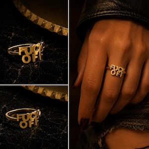 May include: A gold-toned metal ring featuring the text FUCK OFF as the centrepiece. The letters are integrated into the band design, creating a bold statement piece. The ring is shown both as a standalone item and worn on a finger.