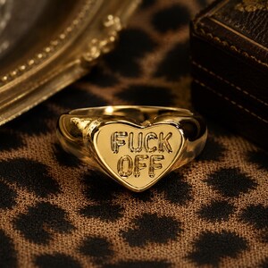 May include: A gold-toned heart-shaped ring featuring the engraved text FUCK OFF in capital letters on the front surface. The ring has a smooth, polished finish and a rounded band, showcasing a bold design with clear, legible lettering.