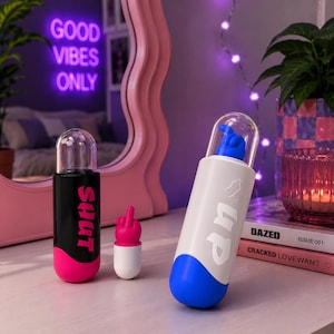 May include: Two cylindrical containers and a small figurine. One black container features the pink text SHUT and a pink base. One white container features the white text up and a blue base. A small pink figurine shows a middle finger gesture.