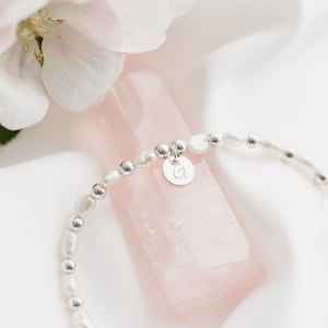 May include: A delicate bracelet made with small white pearls and silver beads. The bracelet has a silver charm with the letter "G" engraved on it.