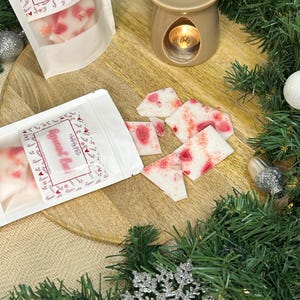 Op de afbeelding: Twee witte zakjes met rode en witte ontwerpen die pepermunt-wasmelts bevatten. De zakjes liggen op een houten oppervlak met een bruine, keramische waswarmer en een brandende kaars erin. De achtergrond toont een groene krans met een zilveren sneeuwvlok-versiering.