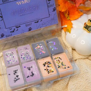 May include: A clear plastic box containing six small wax melts. The wax melts are in various colours and have glitter and small black bats on top. The box has a purple label that says "WWPMC HALLOWEEN COLLECTION BOX COCONUT & RAPESEED WAX MELT BOO!" with bats and a spider web design.