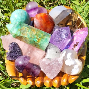 May include: A wooden bowl filled with various crystals and gemstones, including amethyst, rose quartz, agate, and green aventurine. The bowl is surrounded by green grass.