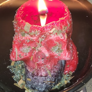 May include: A red skull-shaped candle with a lit wick, surrounded by dried herbs and crystals in a black bowl.