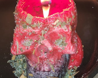 May include: A red skull-shaped candle with a lit wick, surrounded by dried herbs and crystals in a black bowl.