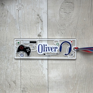 May include: A clear plastic bookmark with a blue and white design. The bookmark features a video game controller, headphones, and the name "Oliver".