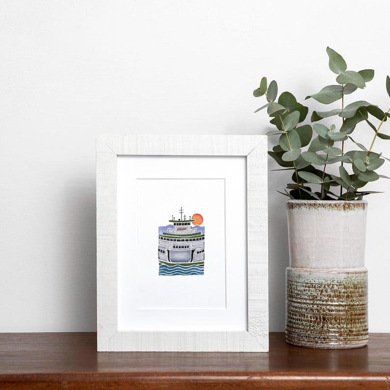 May include: A framed watercolor print of a ferry boat with a red sun in the background. The ferry is white, blue, and green, with blue water at the bottom. The frame is white, and the print is on a wooden surface.