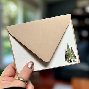 May include: A small, cream-colored greeting card with a tan envelope. The card features a green and gold tree design in the lower right corner. The envelope is closed, and the card is held in a hand.
