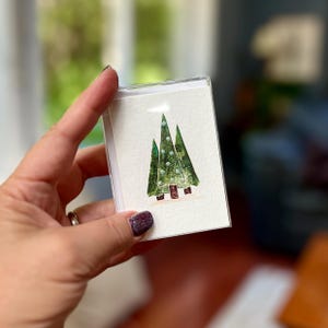 May include: A small, white card featuring a watercolor painting of three green Christmas trees with white dots and brown bases. The card is held in a clear plastic case. The image is in focus.