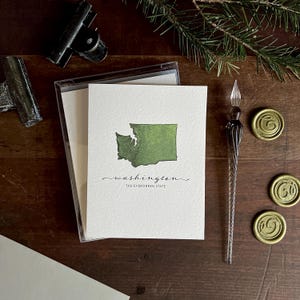 May include: A set of white cards featuring a watercolor map of Washington state, with the word "washington" and the phrase "THE EVERGREEN STATE" in script. The cards are in a clear plastic case, with a glass dip pen and gold wax seals.
