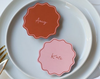 May include: Two place cards with names "Amy" and "Kate" written in white script on a white plate with a gold fork and knife. The place cards are red and pink with scalloped edges.