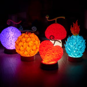May include: A selection of illuminated, colourful fruit-shaped decorative lights. The lights are in various colours, including purple, orange, red, and blue, and are designed to resemble fictional objects. Each light sits on a small, dark base.