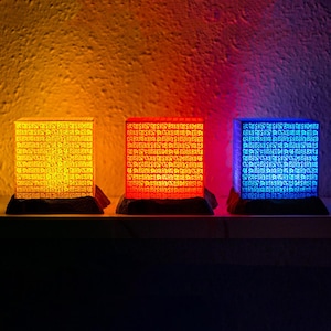 Tres poneglyphs iluminados en tonos naranja, rojo y azul descansan sobre una repisa, proyectando un aura mística y ancestral en la habitación