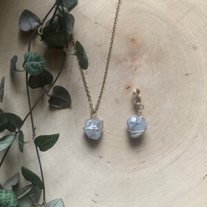 K&ouml;nnte beinhalten: Eine goldfarbene Halskette und ein Anh&auml;nger mit einem hellblauen Stein, der mit goldenem Draht umwickelt ist. Die Halskette und der Anh&auml;nger werden auf einer hellbraunen Holzoberfl&auml;che mit gr&uuml;nen Blattranken im Hintergrund pr&auml;sentiert. Handgefertigter Schmuck.