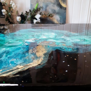 Puede incluir: Primer plano de una mesa de resina negra con un dise&ntilde;o abstracto azul, verde y dorado. El dise&ntilde;o se asemeja a un oc&eacute;ano arremolinado con acentos dorados.
