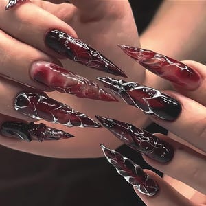 Puede incluir: Un conjunto de uñas artificiales largas y puntiagudas. Las uñas tienen un degradado de rojo oscuro a negro, con diseños blancos en forma de telaraña. Algunas uñas tienen un aspecto texturizado en 3D.