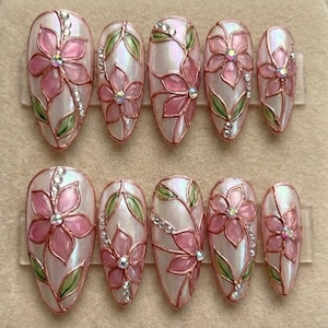May include: A set of ten artificial nails with a pearlescent finish, decorated with pink floral designs, green leaves, and rhinestone accents. The oval-shaped nails are arranged in two rows of five.