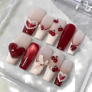 Pode incluir: Um conjunto de unhas artificiais com vários designs. Algumas são vermelhas sólidas, outras rosa claro com detalhes vermelhos, cerejas, corações e enfeites de pérolas. As unhas estão dispostas sobre uma superfície transparente.