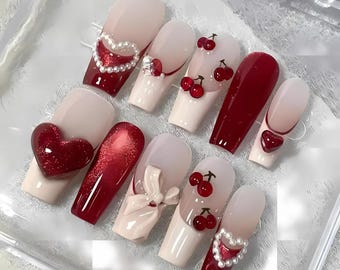 May include: A set of artificial nails in various designs. Some nails are solid red, others are light pink with red accents, cherries, hearts, and pearl embellishments. The nails are arranged on a clear surface.