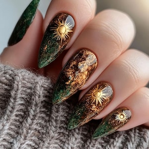 May include: Close-up of a hand with long, almond-shaped nails. The nails feature a gradient design, transitioning from a deep green at the tips to a shimmering brown base. Each nail is adorned with a gold sunburst design.