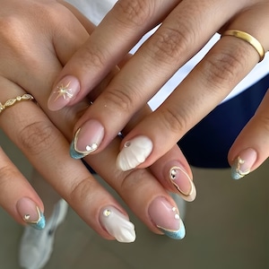 May include: Close-up of manicured nails with a light pink base, adorned with gold accents, rhinestones, and a starburst design. Some nails feature a white, textured design, while others have a light blue French tip with gold trim. A gold ring is visible.