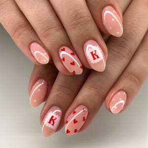 May include: Almond-shaped nails painted in a pale pink. Some nails feature small red hearts, white dots, or a white design with the letter "K" in red. The nails are well-manicured and showcase a Valentine's Day theme.