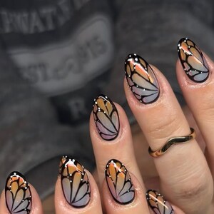 May include: Oval nails with a butterfly wing design. The nails display a gradient of orange, pink, and grey, outlined in black. Small white dots are placed along the wing pattern. A gold ring is visible on one finger.