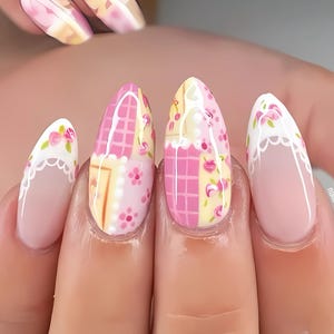 Op de afbeelding: Close-up van gemanicuurde nagels met een lentethema. De nagels hebben een zachtroze basis met ingewikkelde ontwerpen, waaronder bloempatronen, roze en gele ruitpatronen en witte kanten details. De algehele esthetiek is delicaat en vrouwelijk.