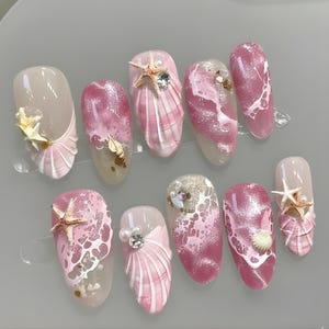May include: A set of ten artificial nails with a beach theme. The nails feature a variety of designs, including pink and white seashell patterns, starfish, and pearl accents. Some nails have a glittery finish.