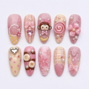 Könnte beinhalten: Ein Set aus zehn mandelförmigen Press-on-Nägeln in Rosa- und Cremetönen. Die Designs umfassen ein Affengesicht, eine Banane, ein rosa Herz, eine Spirale, eine Blume, Schleifen, Perlen, Strasssteine, Punkte, Streifen und ein Gittermuster mit 3D-Akzenten.