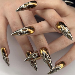 May include: Gold and silver metallic nail art. The long, pointed nails have a gold base with silver flame designs. The nails are a striking accessory, perfect for a bold fashion statement.