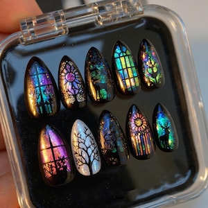 May include: A collection of ten artificial nails with a dark, glossy finish. Each nail showcases unique designs, including stained glass window patterns, a tree, and a deer silhouette, with iridescent colour effects. The nails are almond-shaped.