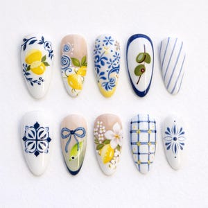 Könnte beinhalten: Zehn mandelförmige Kunstnägel mit handgemalten Designs. Die Designs zeigen Zitronen, Blumenmuster, Oliven und geometrische Formen in Blau, Gelb und Weiß. Die Nägel sind auf einer weißen Oberfläche angeordnet.