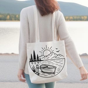 Puede incluir: Un bolso de tela blanco con un gr&aacute;fico de l&iacute;neas negras que muestra un bote de remos en un lago, un poste de madera, hierba alta, pinos, monta&ntilde;as y un sol. El bolso tiene dos correas largas para el hombro y se lleva colgado.