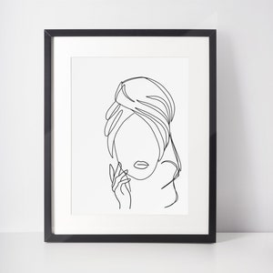 May include: Black and white line art print of a woman's face with her hair pulled back. The print is framed in a black frame.