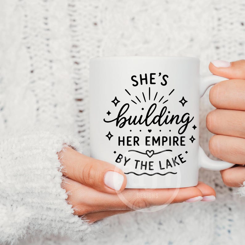 Puede incluir: Una taza de cer&aacute;mica blanca con texto en negro que dice SHE'S building HER EMPIRE BY THE LAKE, decorada con peque&ntilde;os s&iacute;mbolos de estrellas y corazones. El dise&ntilde;o est&aacute; centrado en la parte frontal de la taza, que tiene un asa curva a la derecha.