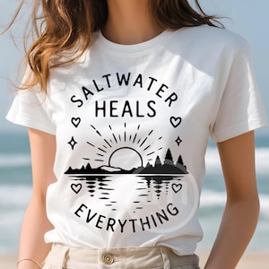 Puede incluir: Una camiseta blanca de manga corta con un estampado gr&aacute;fico negro de un sol saliendo sobre un paisaje, con el texto "SALTWATER HEALS EVERYTHING" arqueado sobre y bajo el dise&ntilde;o, decorado con peque&ntilde;os s&iacute;mbolos de corazones y estrellas.