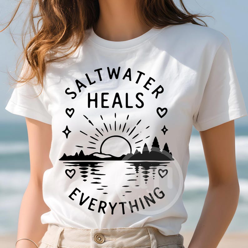 Puede incluir: Una camiseta blanca de manga corta con un estampado gr&aacute;fico negro de un sol saliendo sobre un paisaje, con el texto "SALTWATER HEALS EVERYTHING" arqueado sobre y bajo el dise&ntilde;o, decorado con peque&ntilde;os s&iacute;mbolos de corazones y estrellas.