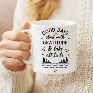 Puede incluir: Una taza de cer&aacute;mica blanca con texto en negro que dice Good days start with GRATITUDE & lake attitude. El dise&ntilde;o incluye peque&ntilde;as ilustraciones de p&aacute;jaros, corazones, estrellas, pinos y l&iacute;neas onduladas de agua en la parte frontal.