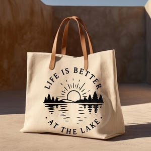 Puede incluir: Un bolso tote de lona beige con dos asas de cuero marr&oacute;n claro. La parte frontal presenta un gr&aacute;fico negro de un sol saliendo sobre un bosque y un lago, con el texto "LIFE IS BETTER AT THE LAKE" dispuesto en un patr&oacute;n circular alrededor.