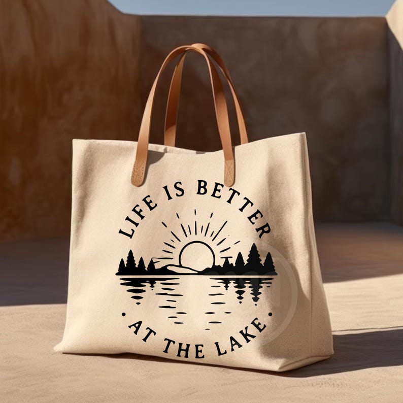 Puede incluir: Un bolso tote de lona beige con dos asas de cuero marr&oacute;n claro. La parte frontal presenta un gr&aacute;fico negro de un sol saliendo sobre un bosque y un lago, con el texto "LIFE IS BETTER AT THE LAKE" dispuesto en un patr&oacute;n circular alrededor.