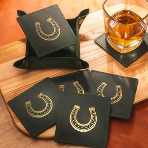 May include: A collection of dark green leather coasters, each featuring a gold horseshoe design. The coasters are square and are presented with a matching tray. A glass of amber liquid rests on one of the coasters.